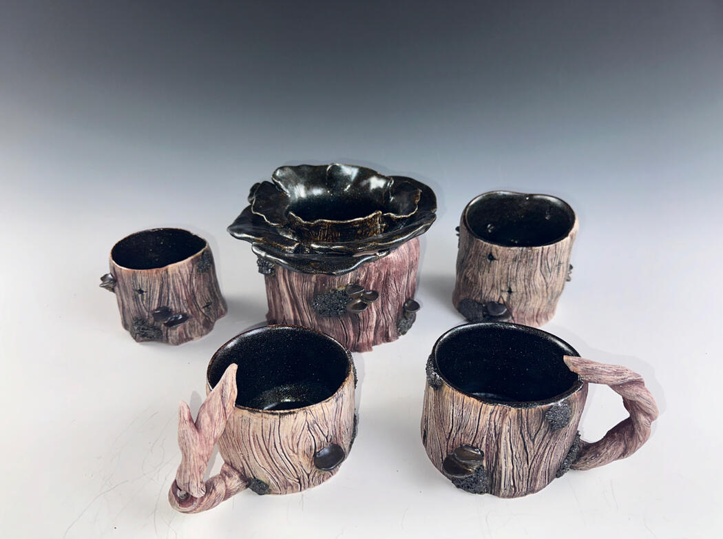 Woodland Tea Party by Carissa Geraud A ceramic drinkware set designed to resemble tree stumps and branches. The set includes cups and a central piece with textured bark-like surfaces, dark interiors, and sculpted details of mushrooms and lichen, evoking a whimsical forest aesthetic.